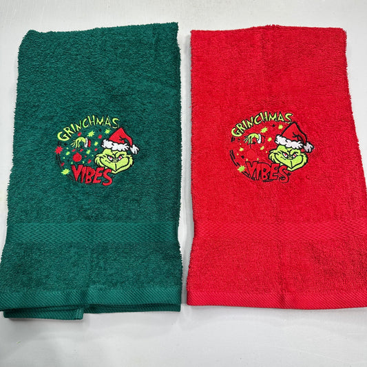 Two folded terry cloth towels side by side on a white background, one dark green and one red, both featuring an embroidered Grinch-themed holiday design with the words 'Grinchmas Vibes'.