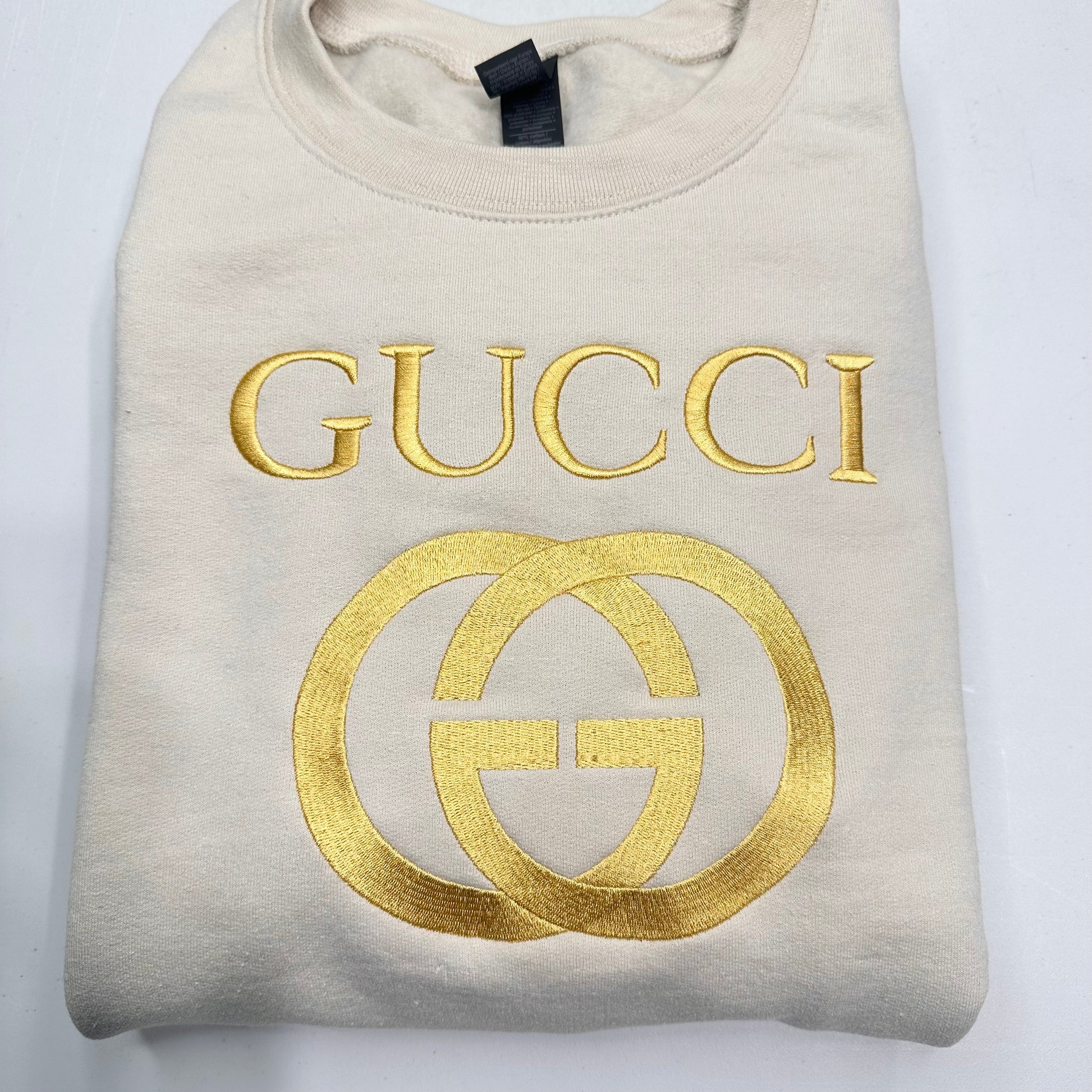 Folded beige sweatshirt with gold Gucci logo on a white background