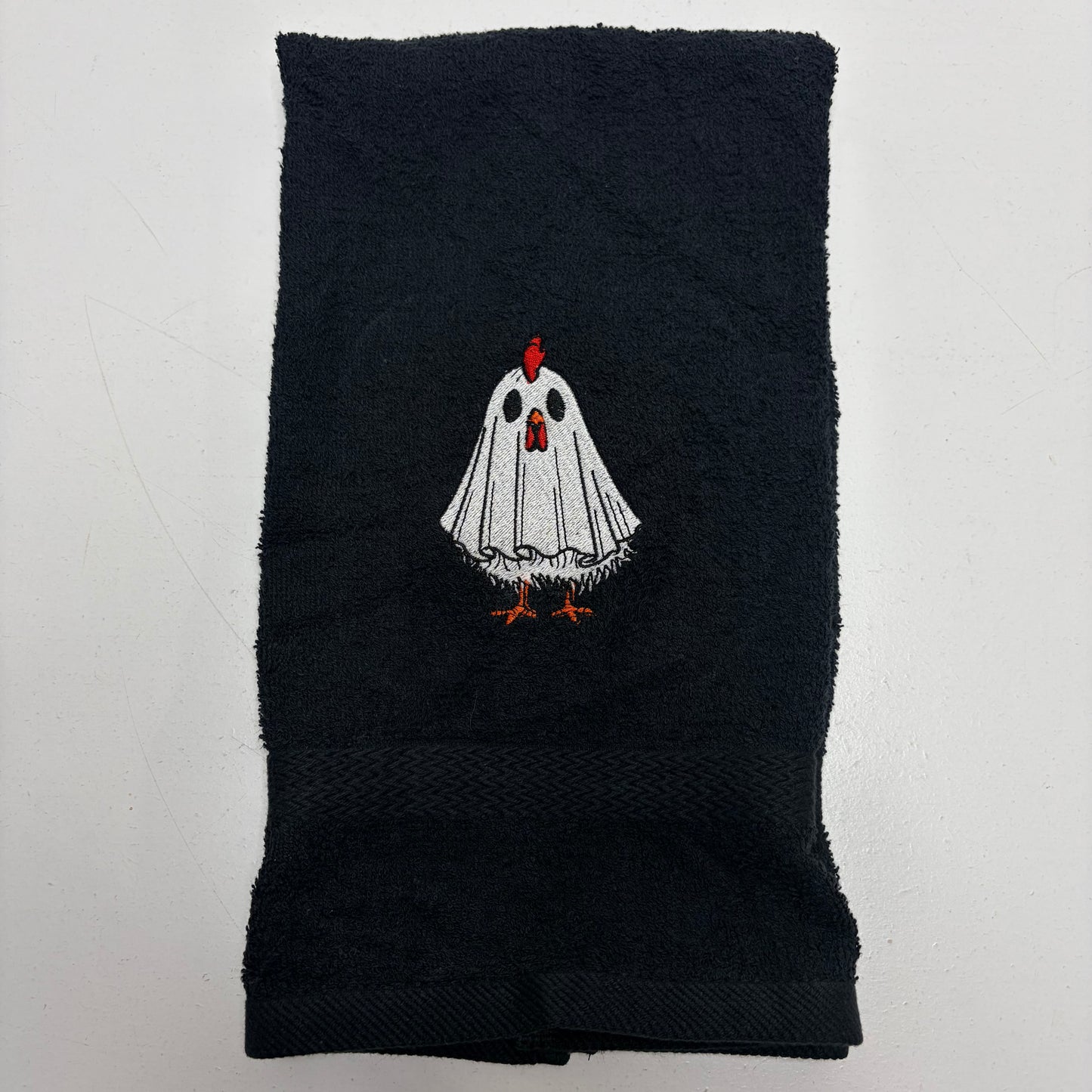 A black cotton towel featuring an embroidered cartoon ghost chicken in the center. The ghost has a white body, black eyes, a red comb, and orange feet.