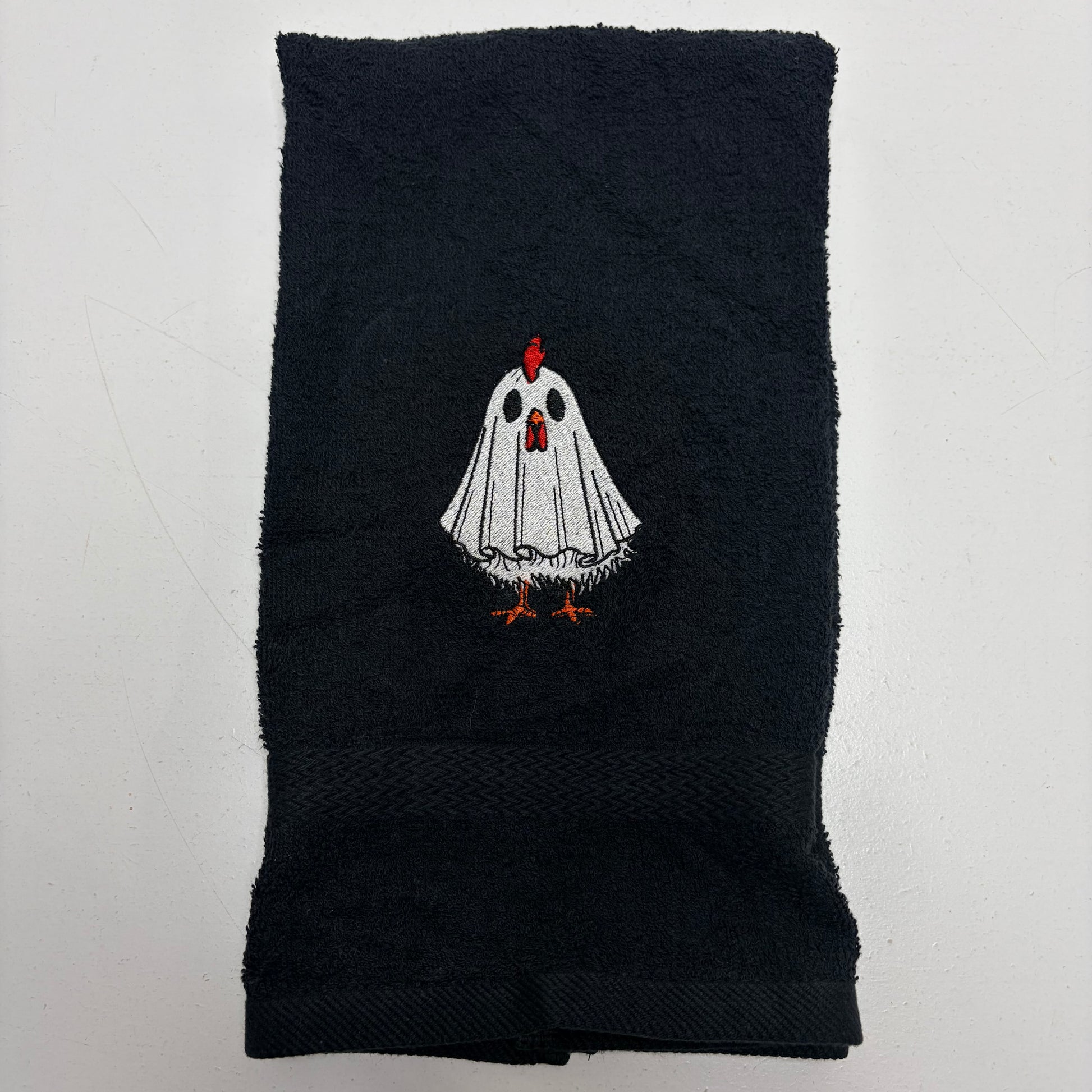 A black cotton towel featuring an embroidered cartoon ghost chicken in the center. The ghost has a white body, black eyes, a red comb, and orange feet.