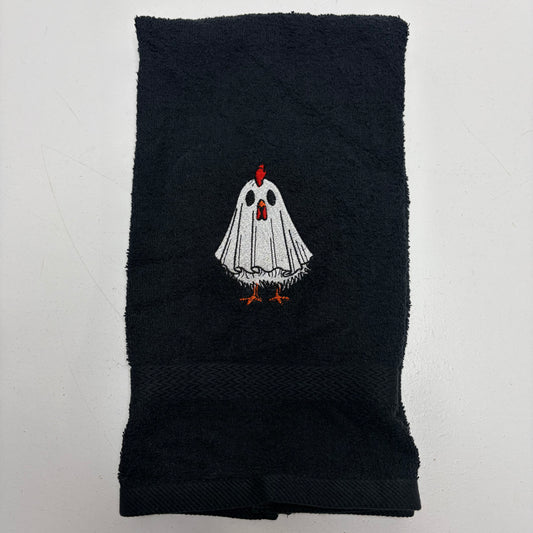 A black cotton towel featuring an embroidered cartoon ghost chicken in the center. The ghost has a white body, black eyes, a red comb, and orange feet.