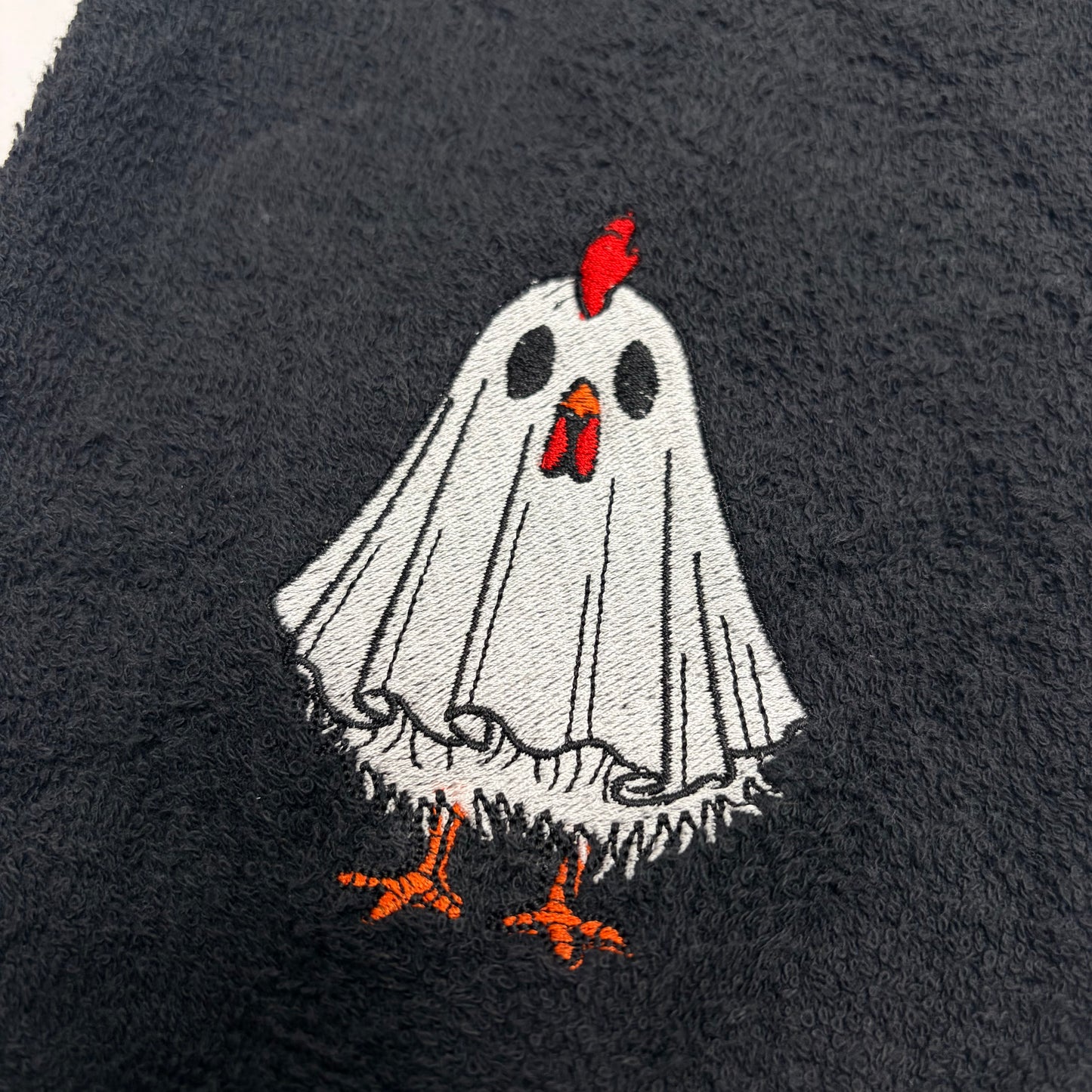 A close-up of a black textured fabric featuring an embroidered cartoon ghost chicken. The ghost has a white body, black eyes, a red comb, and orange feet and beak details.
