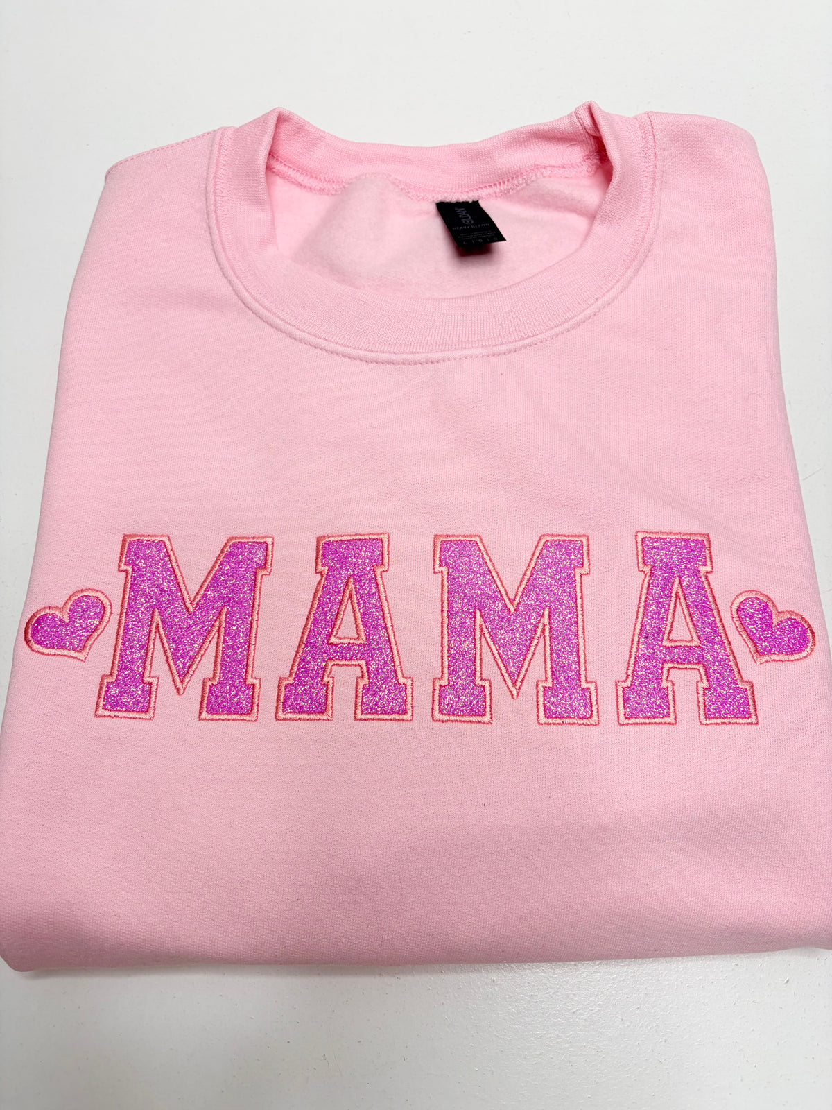 A folded light pink sweatshirt featuring the word 'MAMA' in glittery pink varsity-style block letters outlined in red, flanked by two pink hearts, on a plain white background.