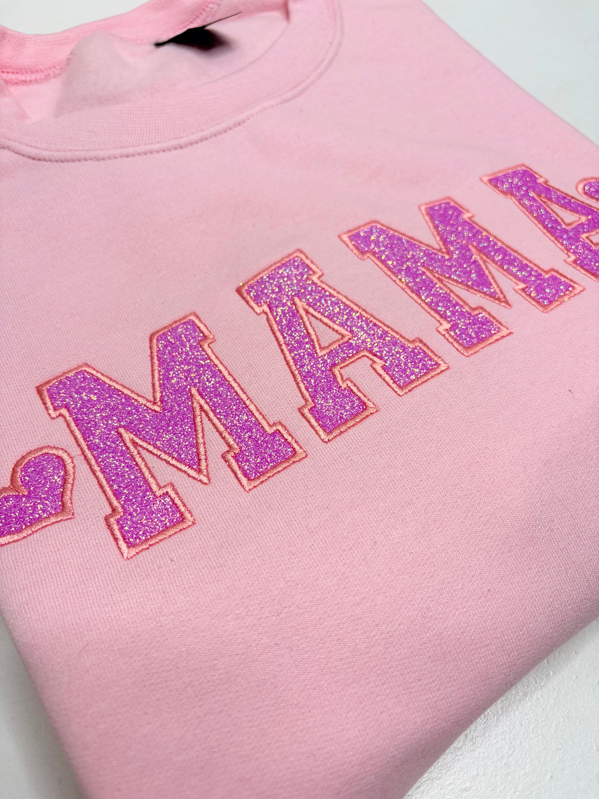 A close-up of a folded light pink sweatshirt featuring the word 'MAMA' in large, glittery purple varsity-style letters with a red outline.