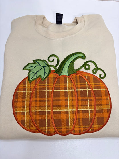 A folded cream-colored sweatshirt featuring a large embroidered orange pumpkin with a plaid pattern, green leaves, and a curled stem on the front.