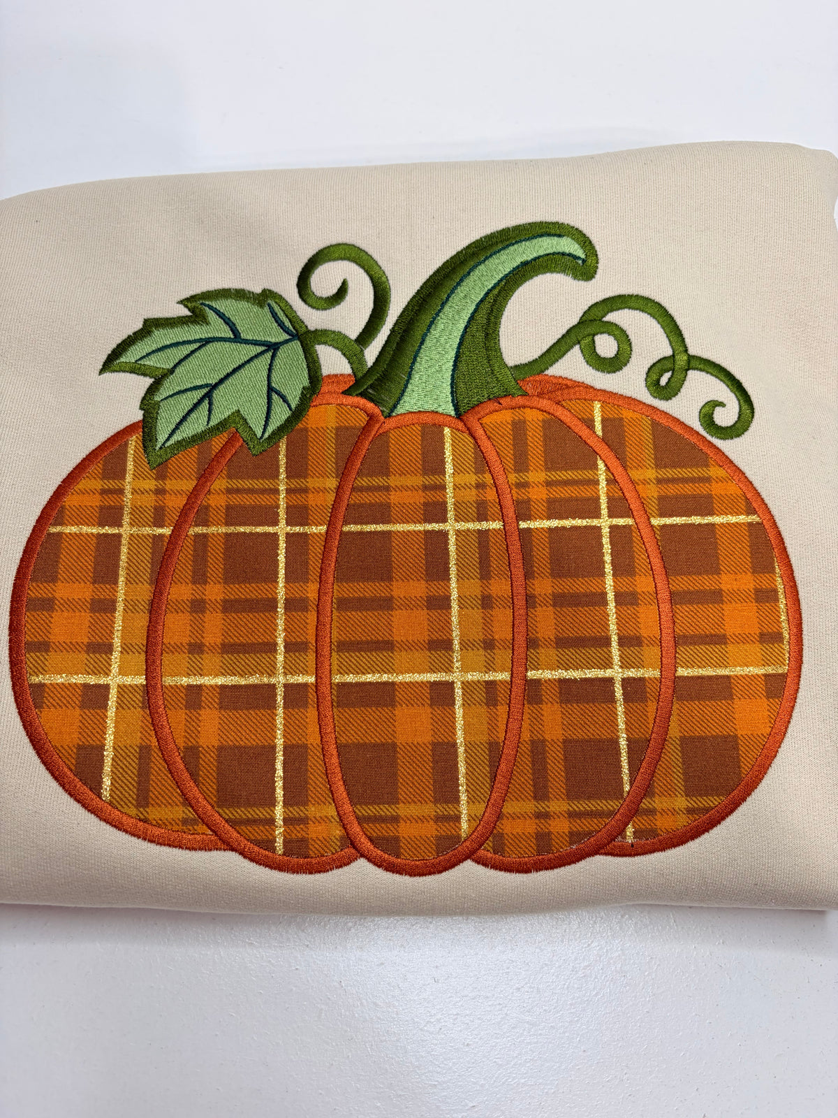 An embroidered pumpkin design on a beige fabric background, featuring an orange and brown plaid pattern with a green stem and leaf.