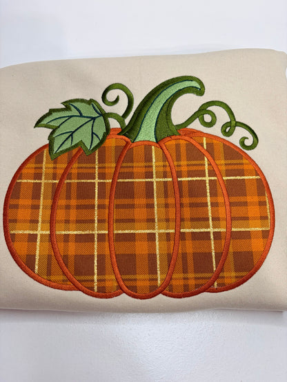 An embroidered pumpkin design on a beige fabric background, featuring an orange and brown plaid pattern with a green stem and leaf.