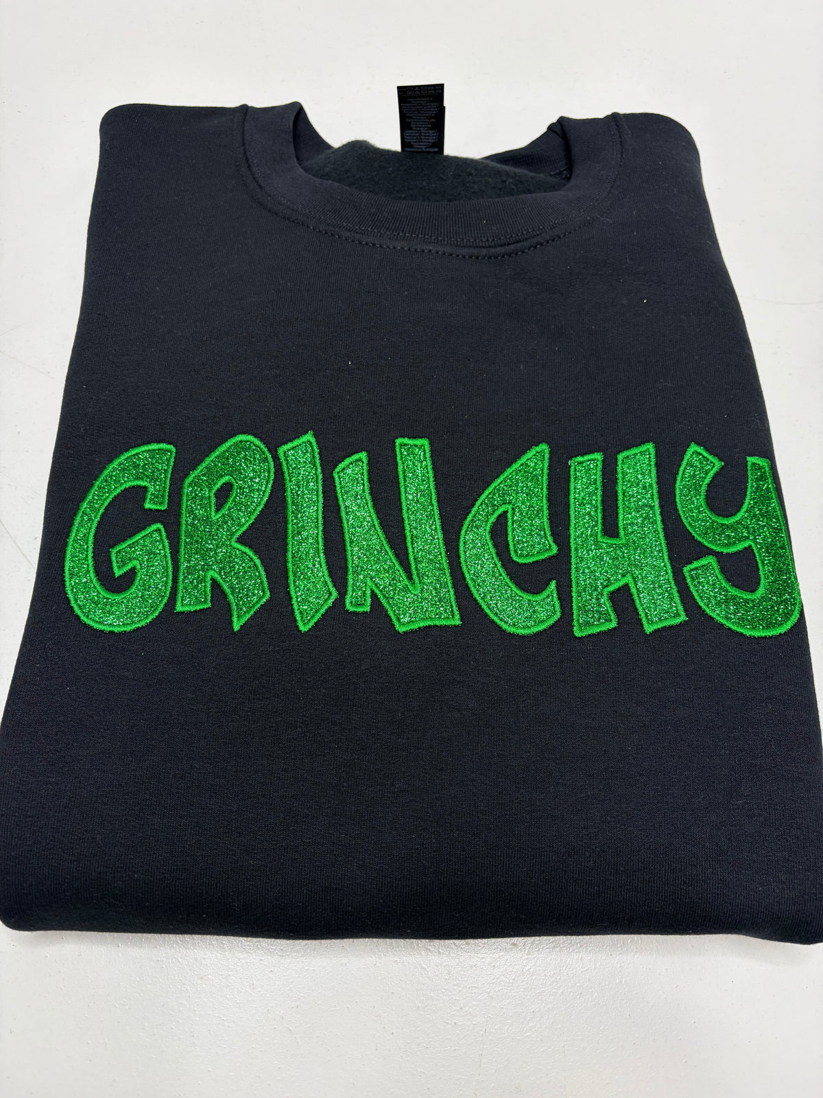 A folded black crewneck sweatshirt featuring the word 'GRINCHY' printed across the chest in a bright green glitter-textured font, displayed on a light gray background.