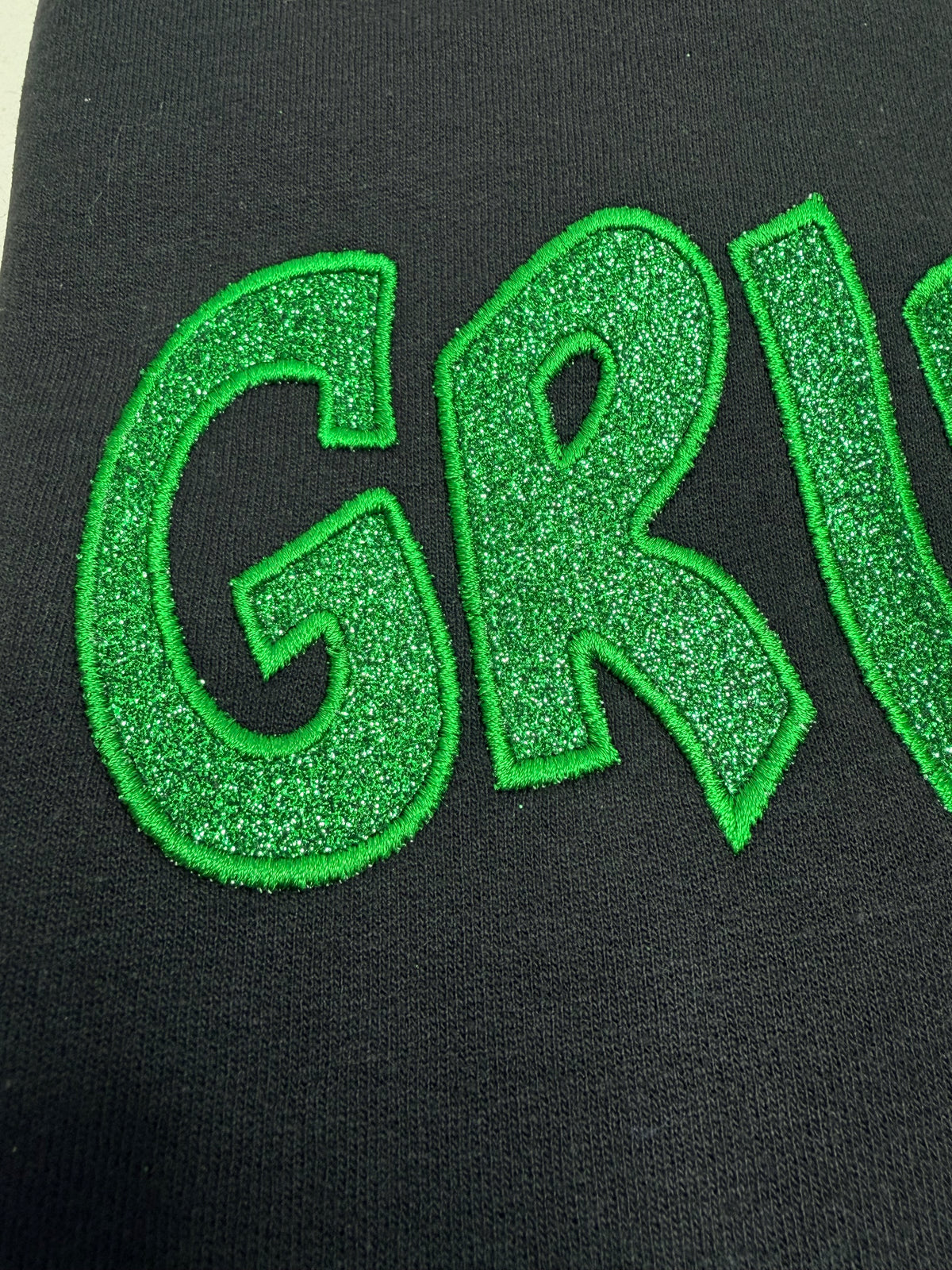 Close-up of a black garment featuring a large, glittery green embroidered letter 'G' and part of the letter 'R' on the front.