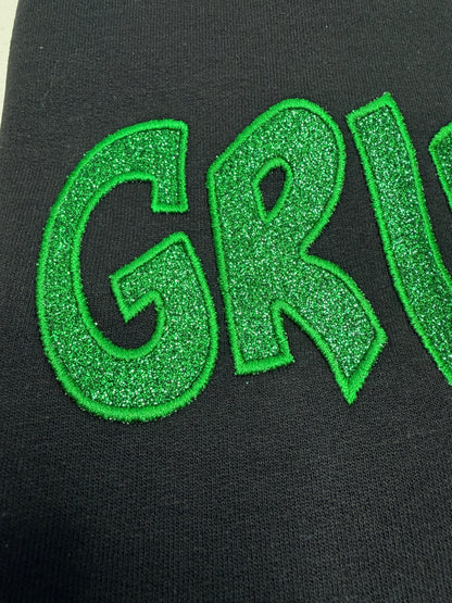 Close-up of a black garment featuring a large, glittery green embroidered letter 'G' and part of the letter 'R' on the front.