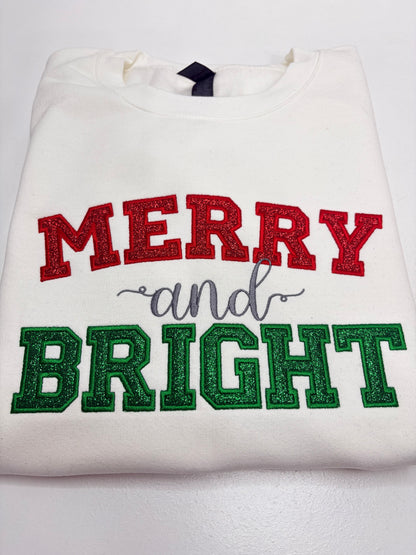 A folded white sweatshirt featuring 'MERRY and BRIGHT' in glittery red and green lettering with a gray script 'and' in the middle, displayed on a white background.