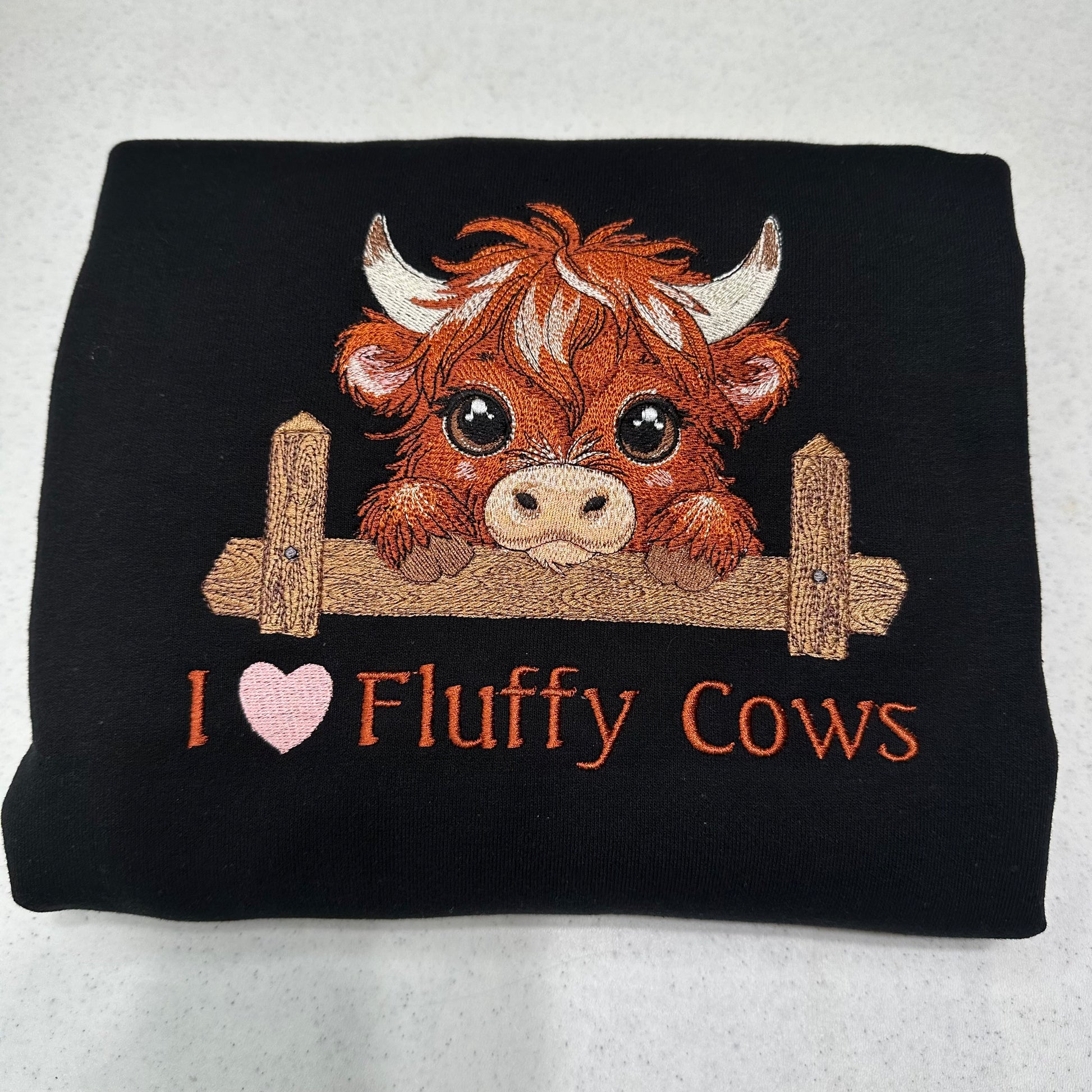 embroidered highland cow with a heart and text on a folded black sweatshirt