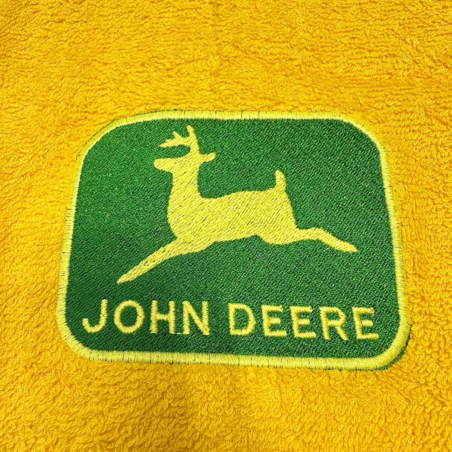 A close-up of a yellow textured fabric, likely a blanket or towel, featuring an embroidered green John Deere logo patch with the classic leaping deer and 'JOHN DEERE' text.