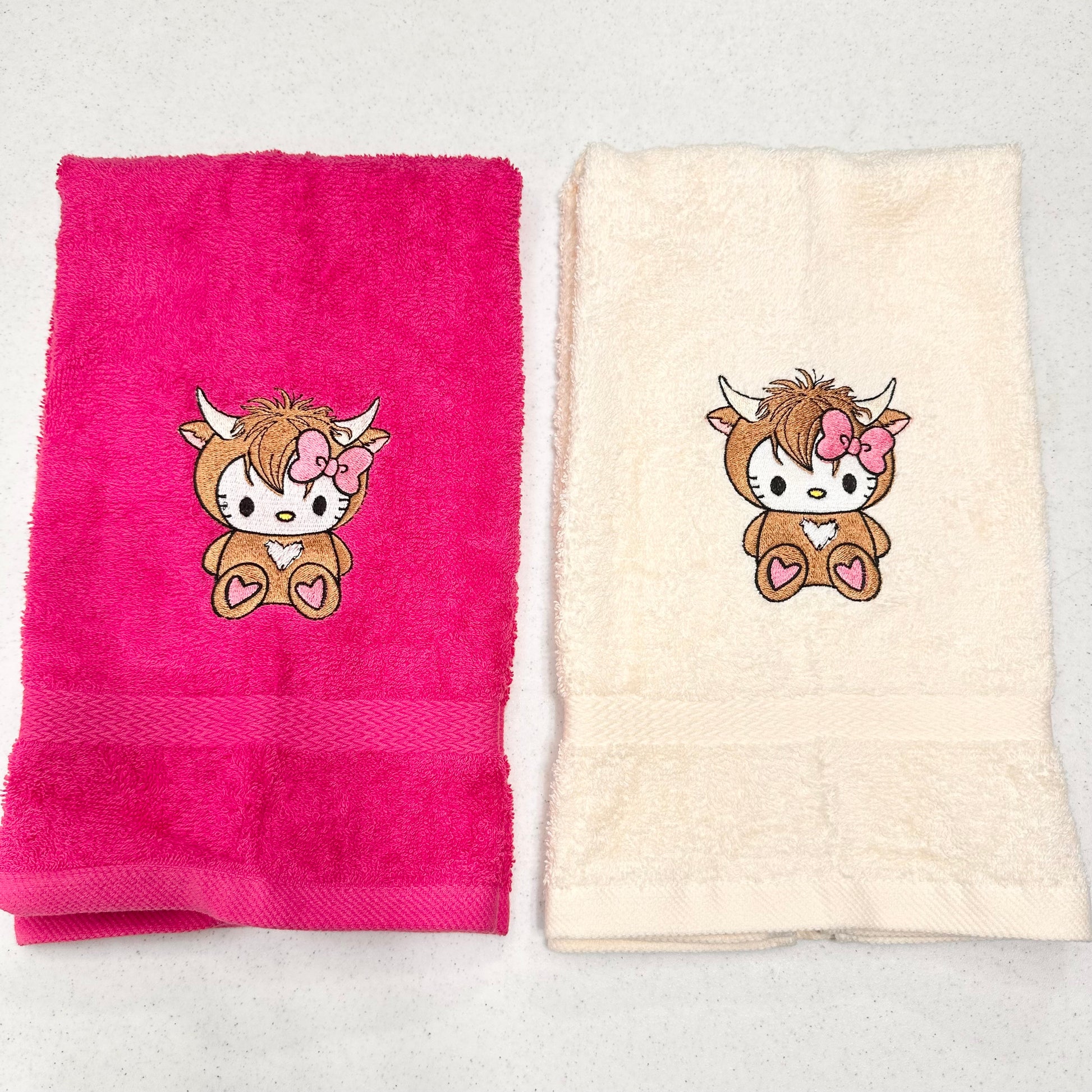 Two folded towels on a light gray surface: one bright pink towel and one cream towel, each featuring a small embroidered cartoon hello kitty highland cow character with a pink bow.