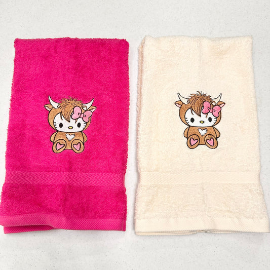 Two folded towels on a light gray surface: one bright pink towel and one cream towel, each featuring a small embroidered cartoon hello kitty highland cow character with a pink bow.