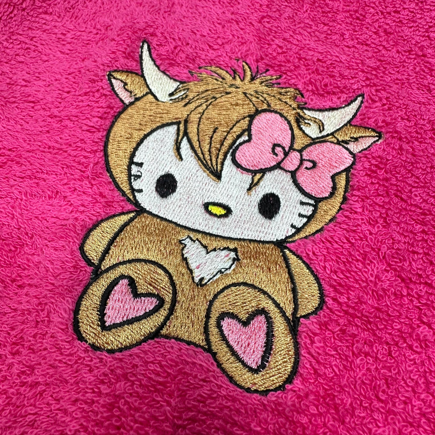 A close-up of an embroidered patch featuring a Hello Kitty-inspired character dressed as a cow, with pink hearts and a pink bow, on a bright pink textured fabric background.