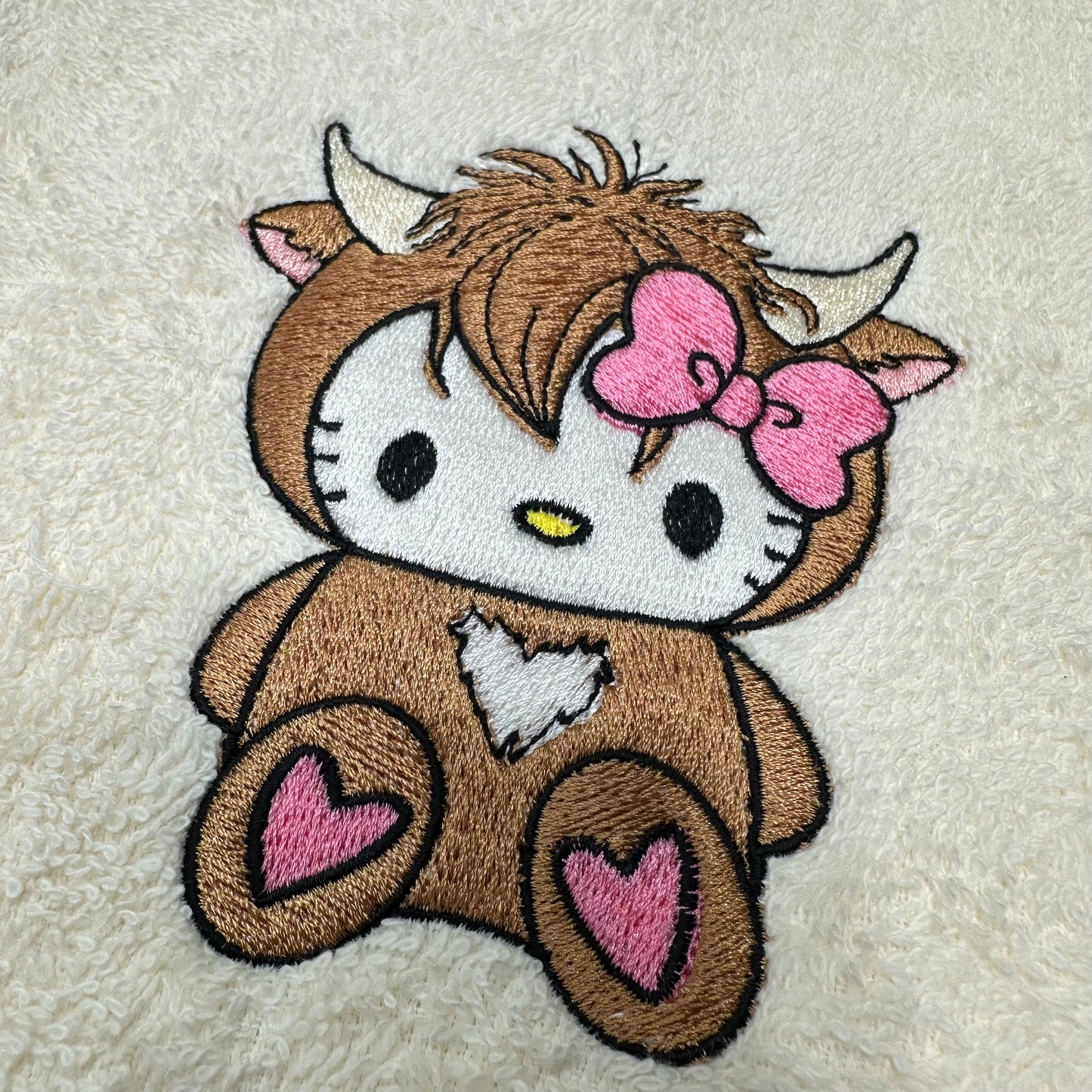 An embroidered patch of a cute cartoon character styled like Hello Kitty in a brown highland cow costume, featuring a pink bow, white face, and pink hearts on the paws, shown on a textured cream background.