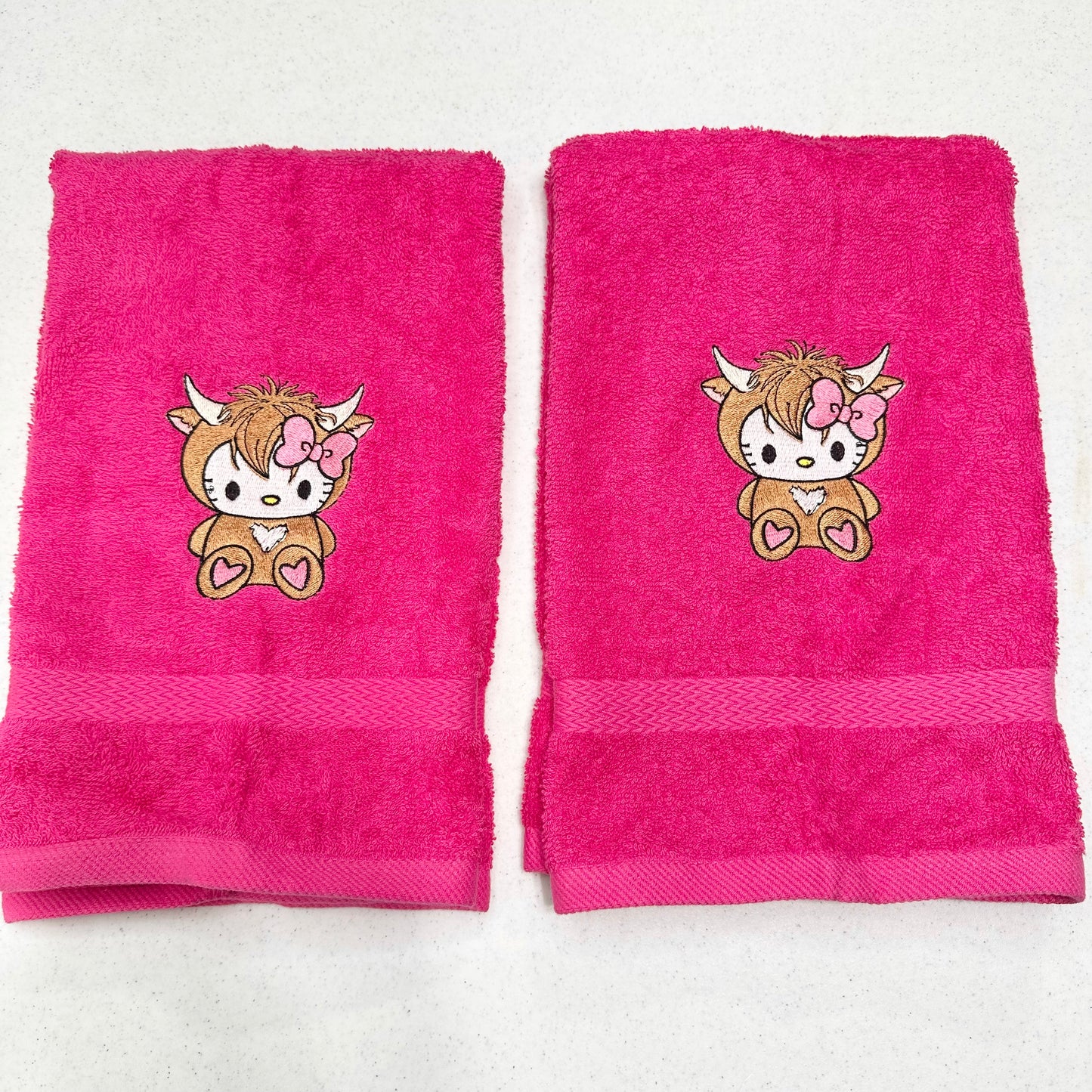 Two bright pink hand towels with embroidered cartoon hello kitty highland cow character motifs, displayed side by side on a light surface.