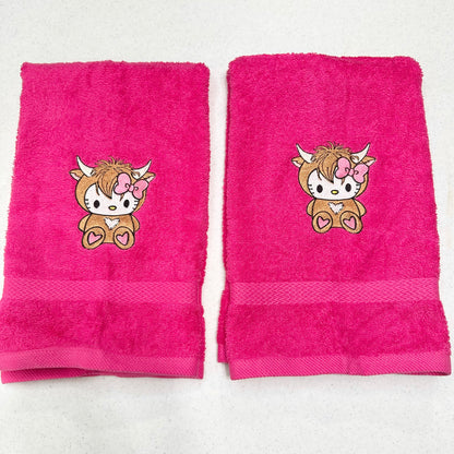 Two bright pink hand towels with embroidered cartoon hello kitty highland cow character motifs, displayed side by side on a light surface.