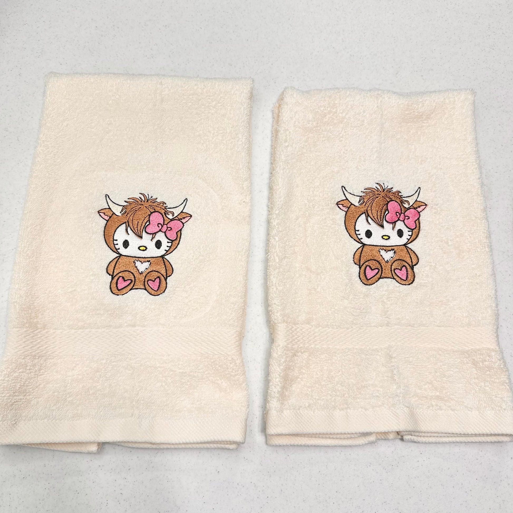 Two cream-colored cotton hand towels laid flat on a light gray surface, each featuring a cartoon hello kitty highland cow character with a pink bow and heart details embroidered on the front.