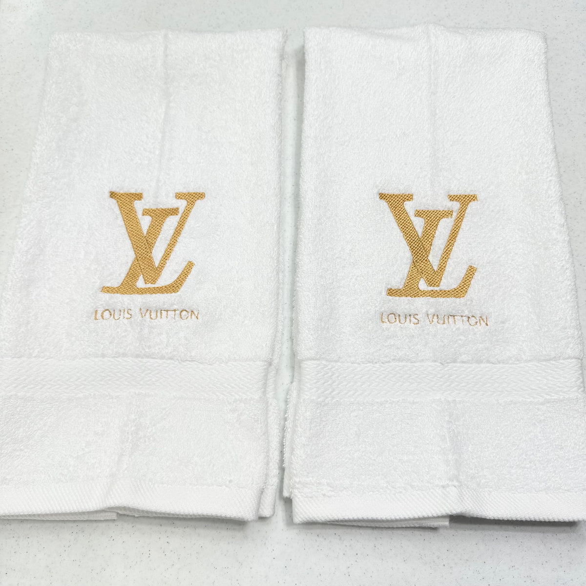 Two white Louis Vuitton bath towels with gold embroidered LV logos and brand names on the front, displayed side by side on a light gray surface.