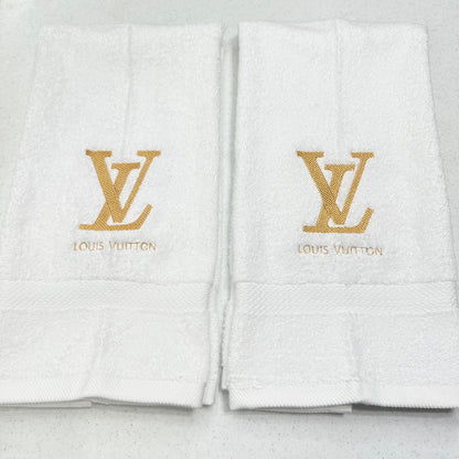 Two white Louis Vuitton bath towels with gold embroidered LV logos and brand names on the front, displayed side by side on a light gray surface.