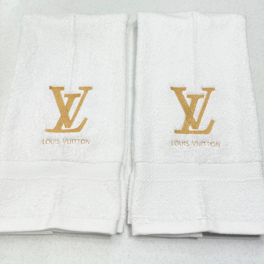 Two white Louis Vuitton bath towels with gold embroidered LV logos and brand names on the front, displayed side by side on a light gray surface.