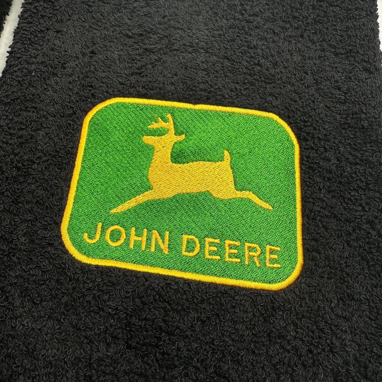 A close-up of a black textured fabric featuring an embroidered John Deere logo patch with a green background, yellow deer silhouette, and yellow text.