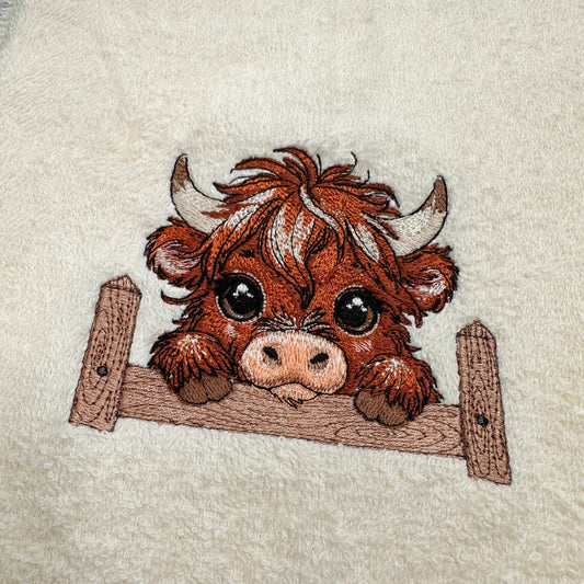 A close-up of a fluffy cream-colored fabric featuring an embroidered cartoon Highland cow peeking over a wooden fence.