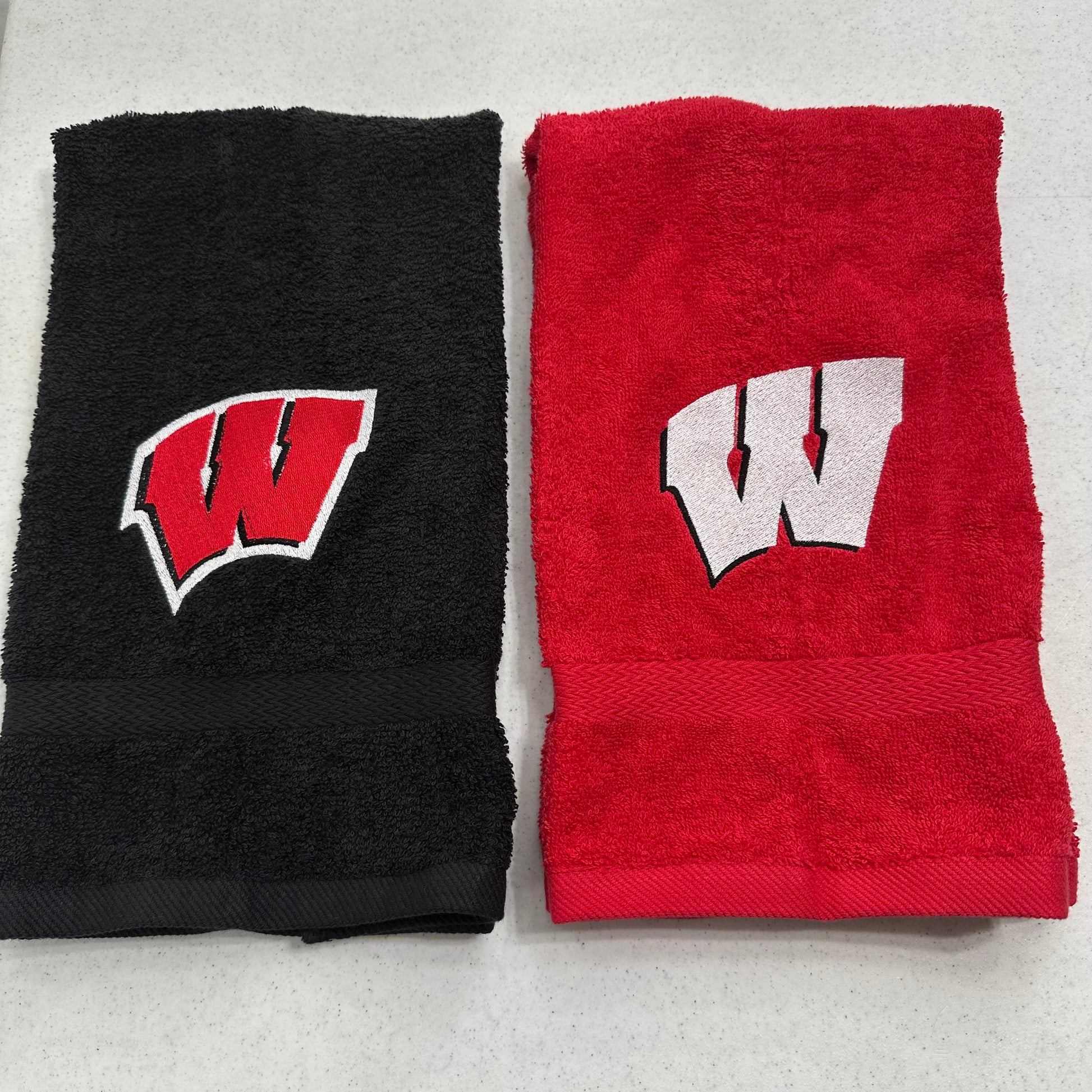 Two folded towels side by side on a light gray surface: one black towel with a red and white embroidered 'W' logo, and one red towel with a white and black embroidered 'W' logo.