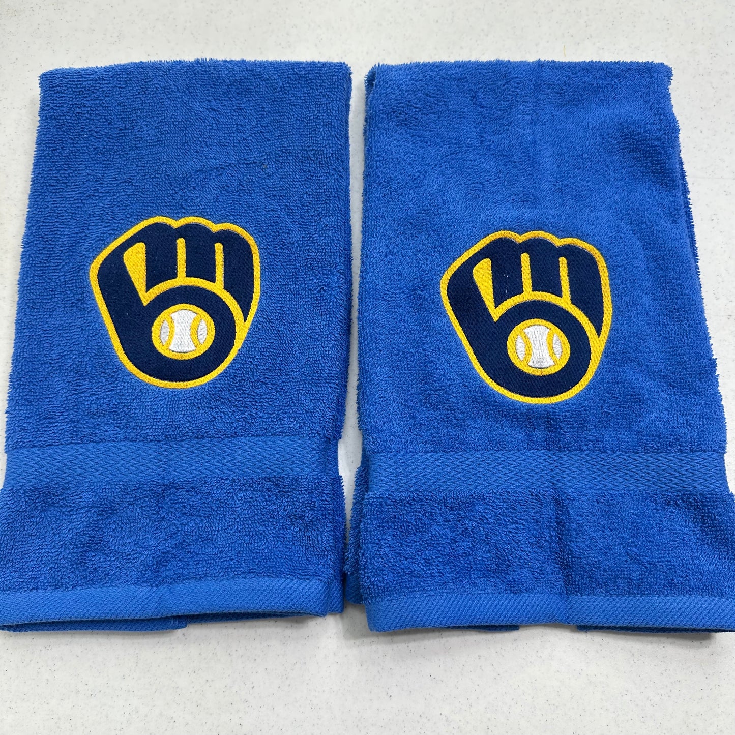 Two blue towels with Milwaukee Brewers baseball glove and ball logos, displayed side-by-side on a light gray surface.