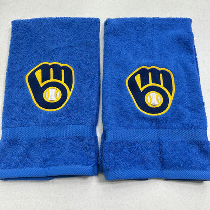Two blue towels with Milwaukee Brewers baseball glove and ball logos, displayed side-by-side on a light gray surface.
