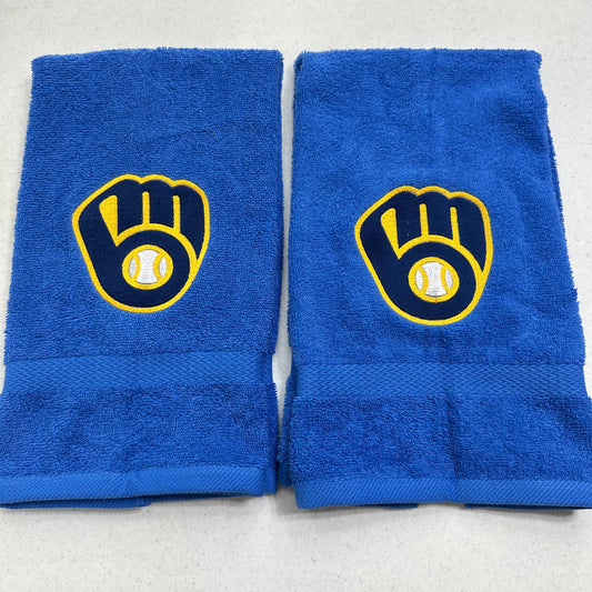 Two blue towels with Milwaukee Brewers baseball glove and ball logos, displayed side-by-side on a light gray surface.