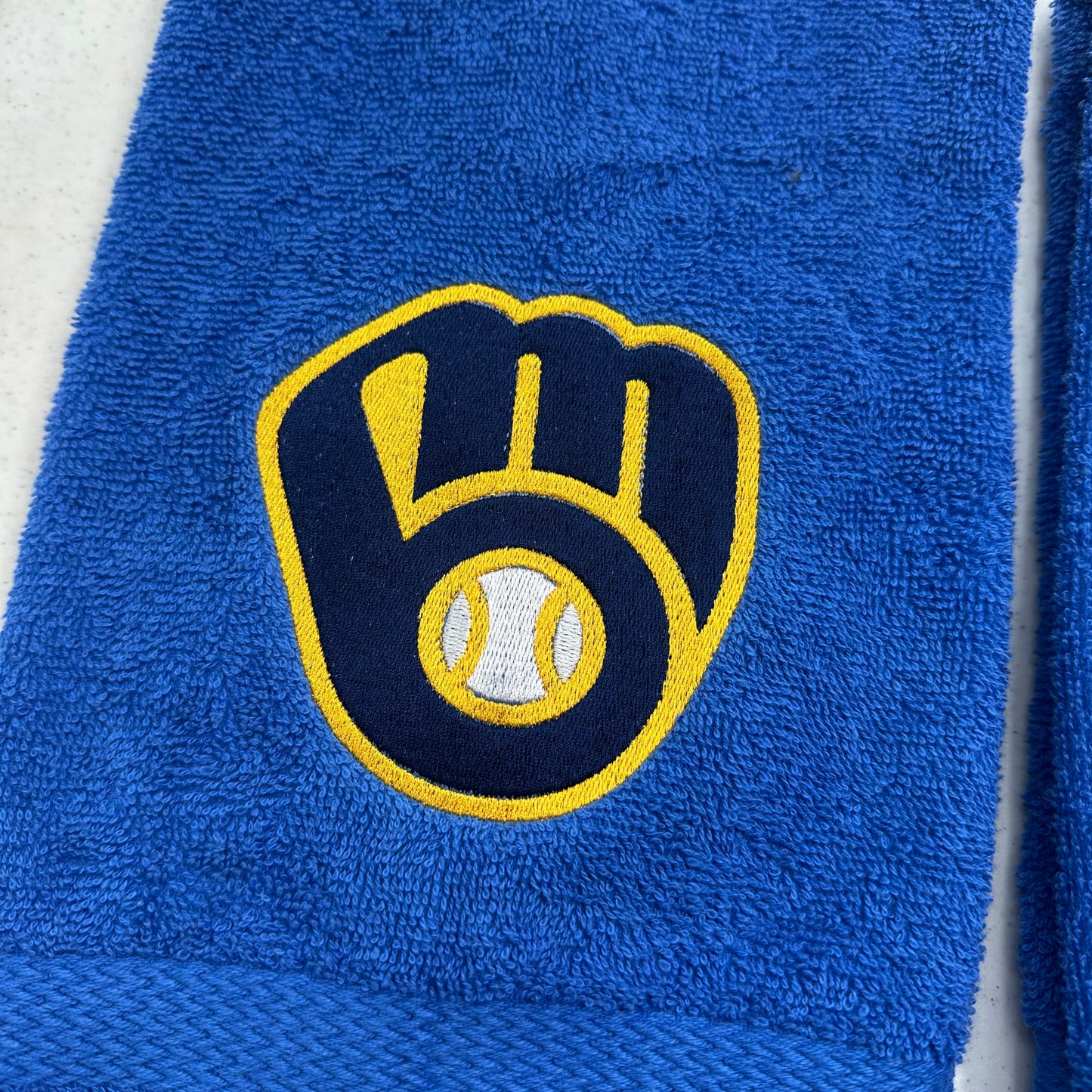 A close-up of a royal blue terry cloth towel featuring an embroidered Milwaukee Brewers logo with a yellow and navy glove and baseball design.