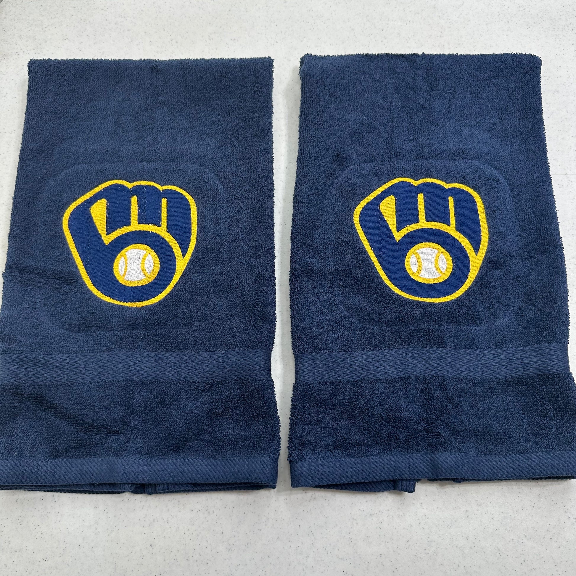 Two navy blue towels with yellow and white Milwaukee Brewers baseball glove and ball logo embroidered on the front, laid flat on a light gray surface.