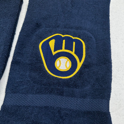 A close-up of a navy blue towel featuring the Milwaukee Brewers baseball glove and ball logo embroidered in yellow and white, with a matching navy woven band near the bottom.