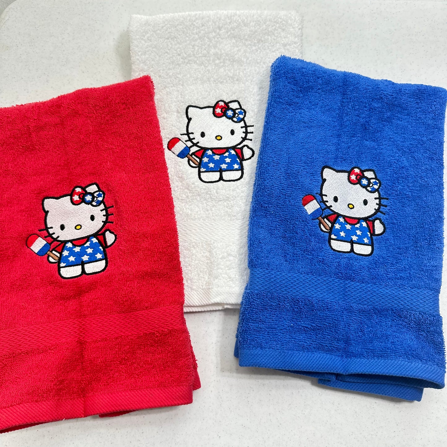 Three folded towels on a light background: a red towel, a white towel, and a blue towel, each embroidered with a Hello Kitty character holding a small American flag.
