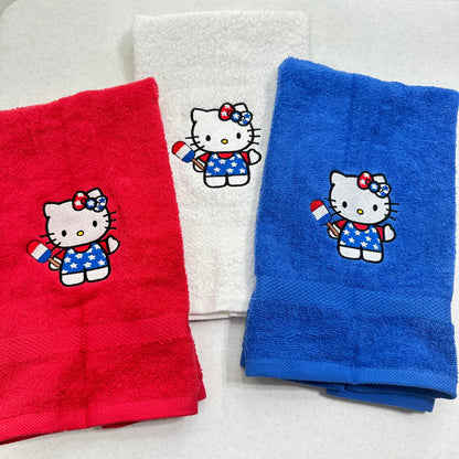 Three folded towels on a light background: a red towel, a white towel, and a blue towel, each embroidered with a Hello Kitty character holding a small American flag.