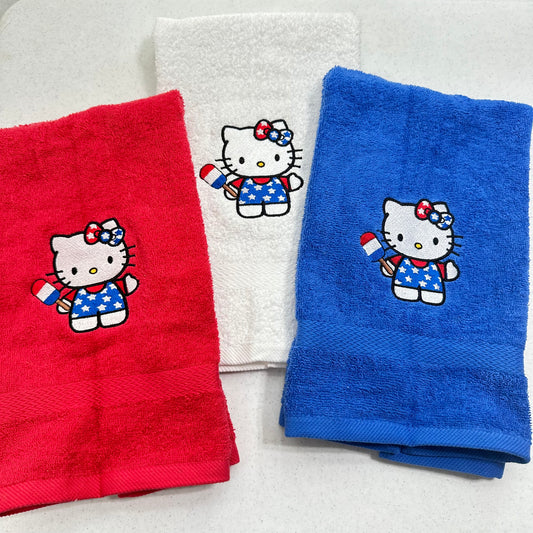 Three folded towels on a light background: a red towel, a white towel, and a blue towel, each embroidered with a Hello Kitty character holding a small American flag.