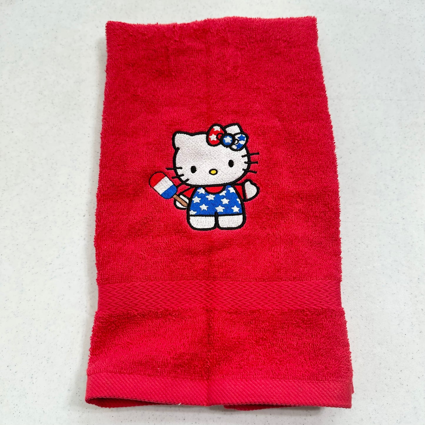 A red terry cloth hand towel featuring an embroidered Hello Kitty character in a patriotic red, white, and blue outfit, displayed flat on a light gray surface.