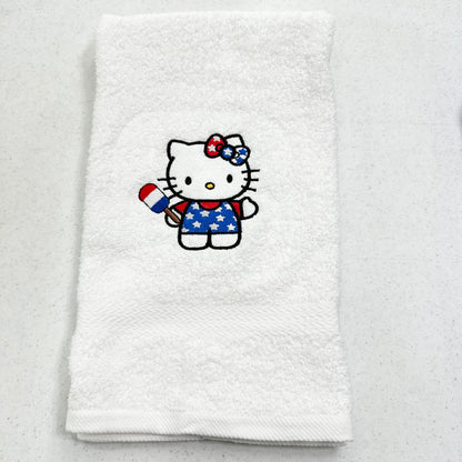A folded white towel featuring an embroidered Hello Kitty character in a patriotic red, white, and blue outfit holding a small flag, set against a light gray speckled background.