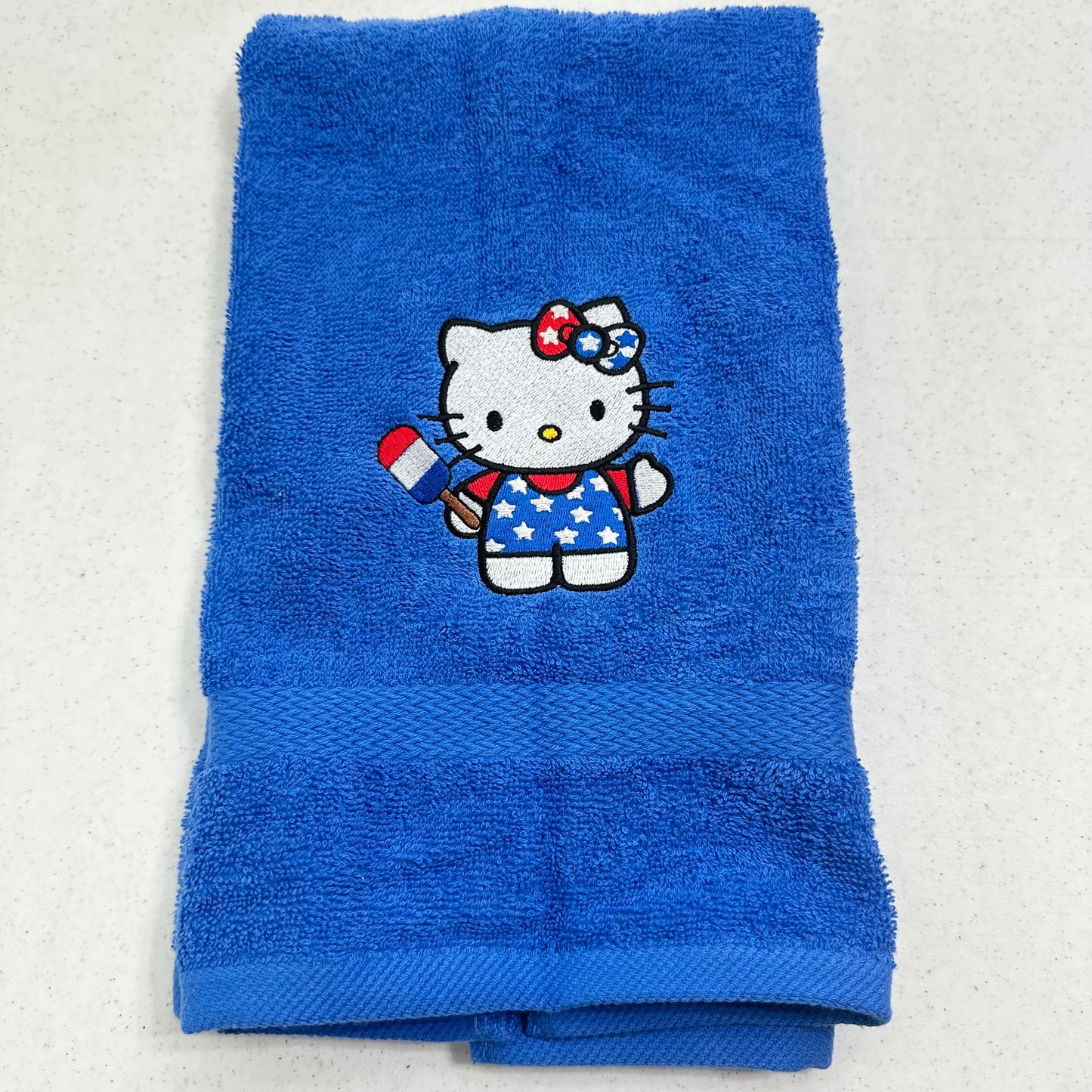 A folded blue towel featuring an embroidered Hello Kitty character holding a red, white, and blue flag, set against a light gray background.