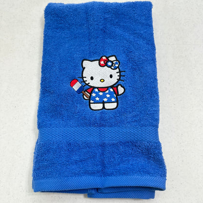A folded blue towel featuring an embroidered Hello Kitty character holding a red, white, and blue flag, set against a light gray background.