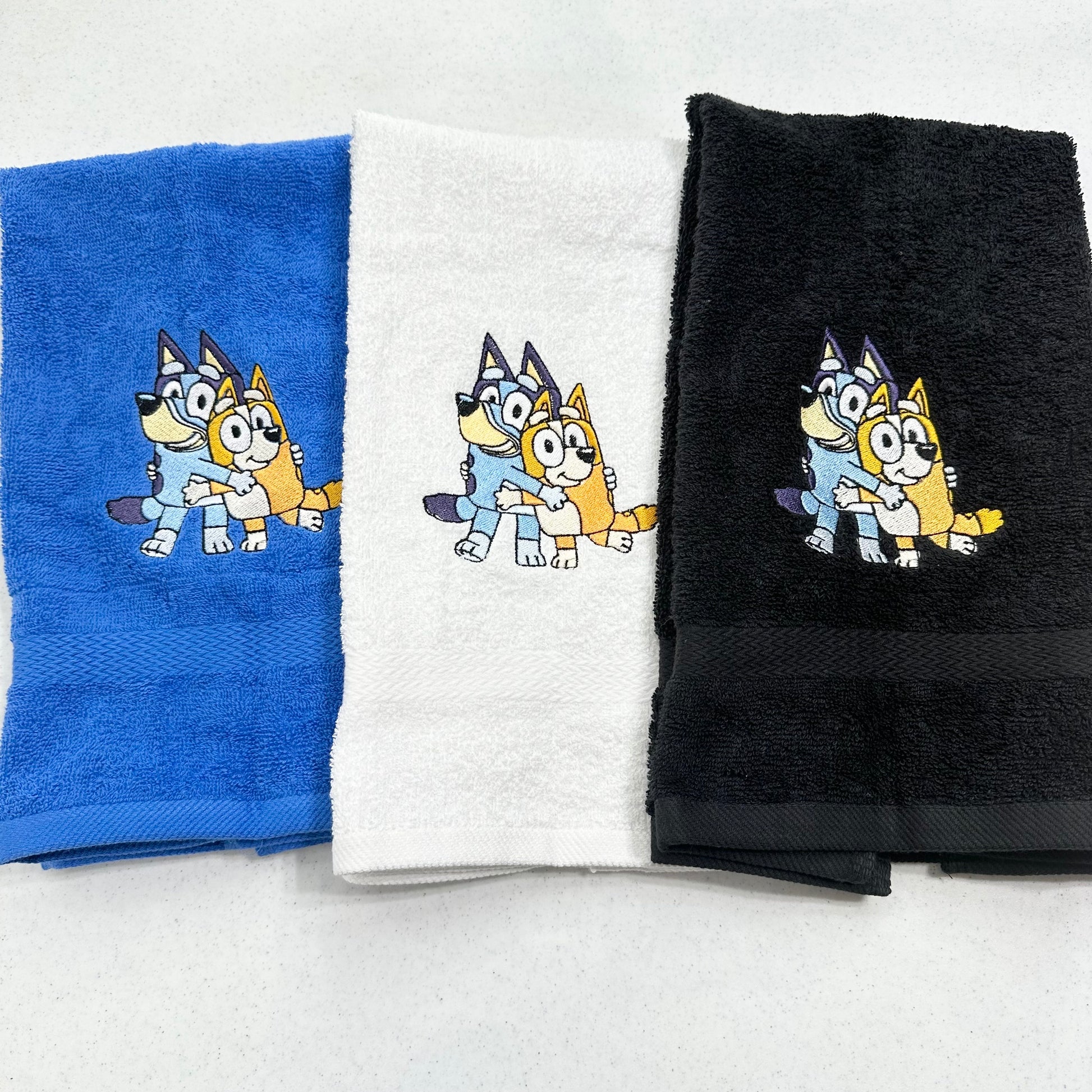 Three folded towels in blue, white, and black, each featuring cartoon bluey and bingo dog characters embroidered near the top center.