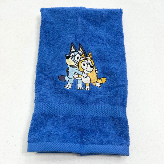 A blue terry cloth hand towel featuring an embroidered cartoon design of Bluey and Bingo hugging in the center, displayed flat against a light off-white background.