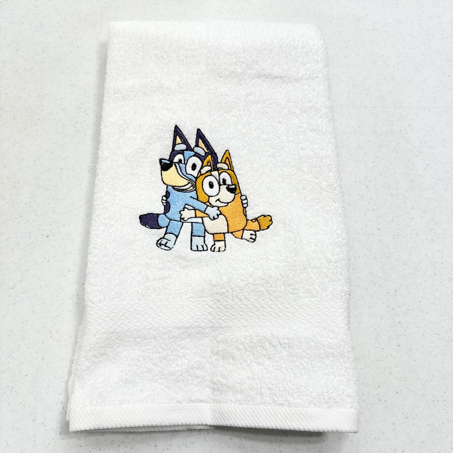 A folded white cotton towel featuring a colorful embroidered illustration of Bluey and Bingo hugging, displayed on a light gray speckled surface.