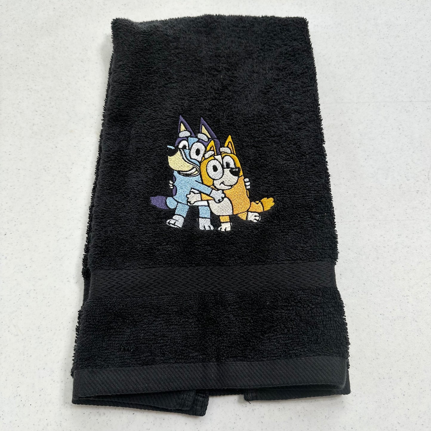 A black terry cloth hand towel featuring an embroidered graphic of Bluey and Bingo from 'The Blue Boy' on the front, displayed flat on a light gray surface.