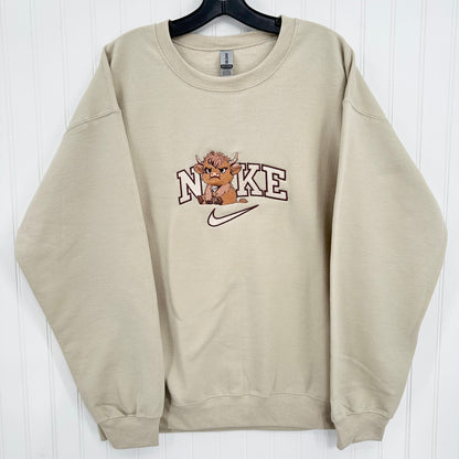 A beige crewneck sweatshirt hanging on a black hanger against a white paneled wall. The front features a graphic with the word 'NIKE' in arched varsity letters, a cartoon highland cow, and a white swoosh.