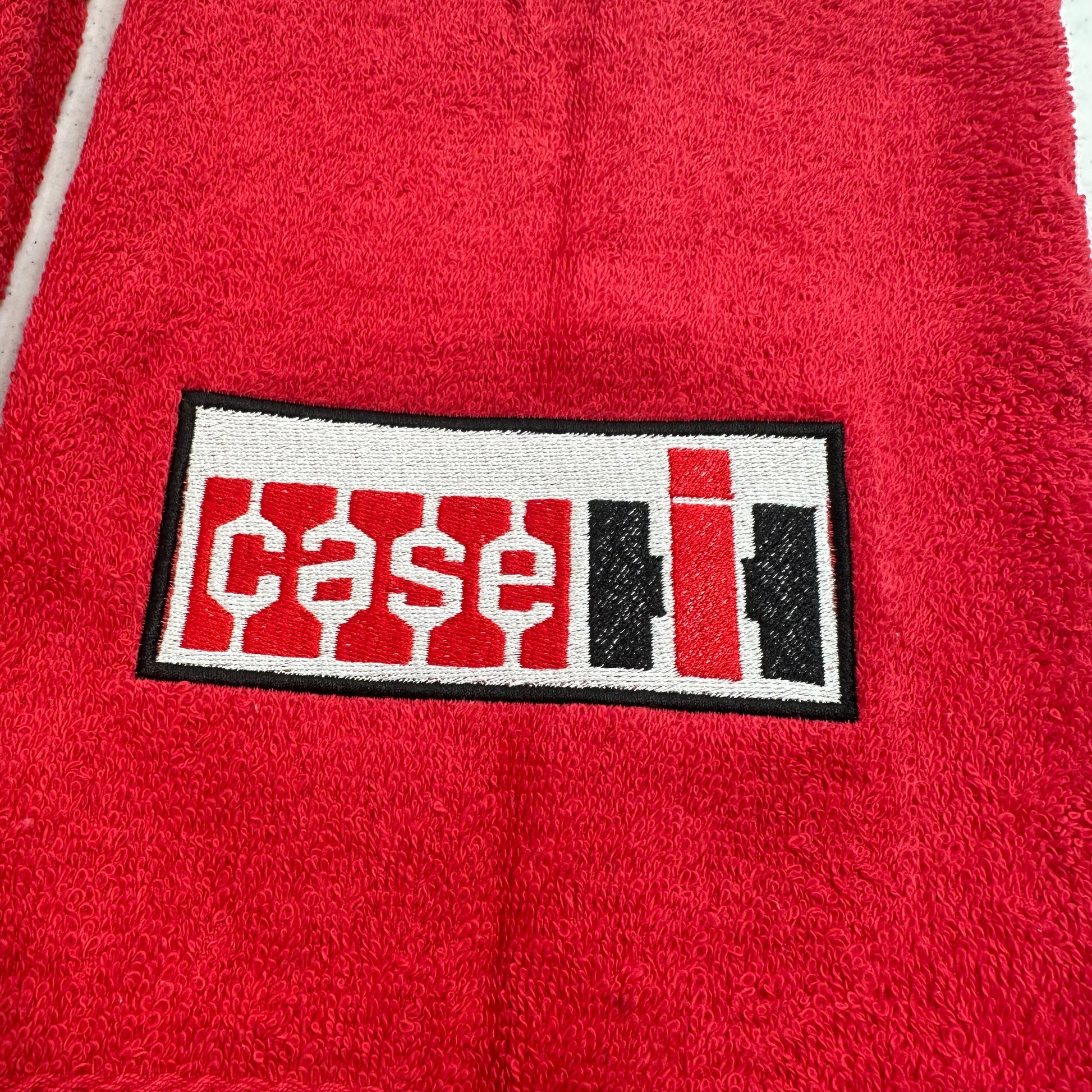 A close-up of a red textured fabric, likely a towel, featuring a rectangular embroidered patch with the word 'case' in white and 'i' in red and black.