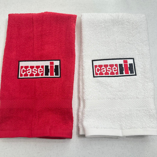 Two folded towels side by side on a light gray surface: a red towel on the left and a white towel on the right, each with a rectangular embroidered patch reading "case" in red, black, and white.
